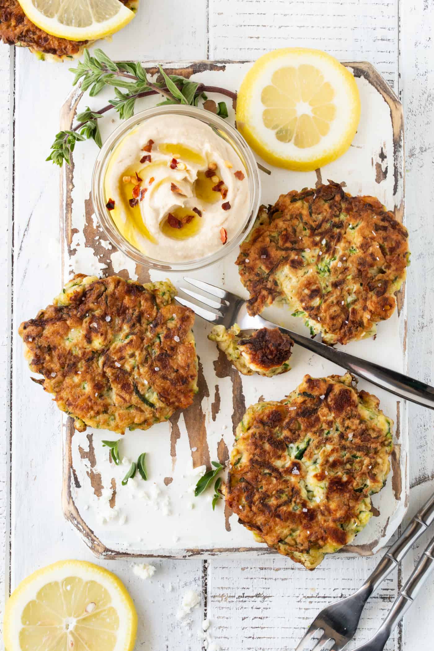 Zucchini Feta Fritters on a white board with a small bowl of hummus. Garnished with fresh oregano and lemon slices.