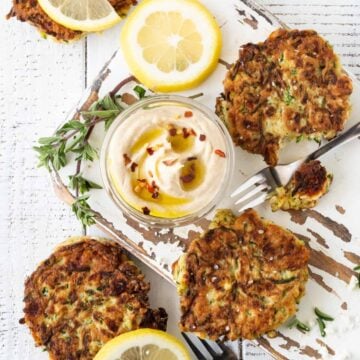 White board filled with zucchini feta fritters. Garnished with fresh hummus, oregano, lemon slices and coarse sea salt.