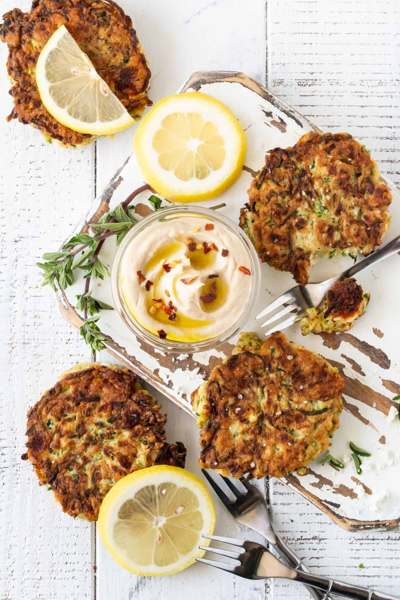 White board filled with zucchini feta fritters. Garnished with fresh hummus, oregano, lemon slices and coarse sea salt.