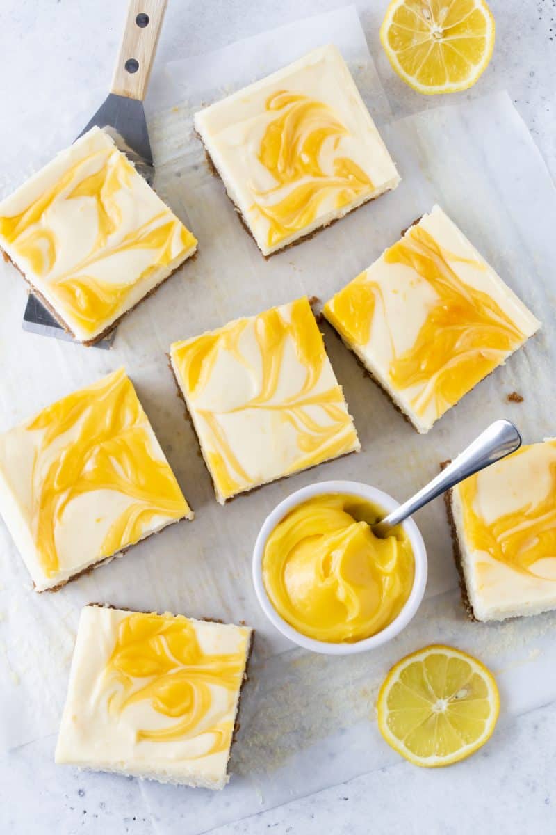 Cheesecake Bars swirled with lemon curd on white parchment paper with a small white bowl of fresh lemon curd. 