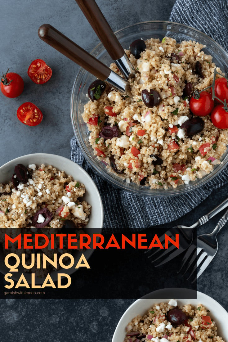 Close up of Mediterranean Quinoa Salad in a glass serving bowl on a dark grey background with wooden spoons for serving. Garnished with fresh cilantro and sliced tomatoes.