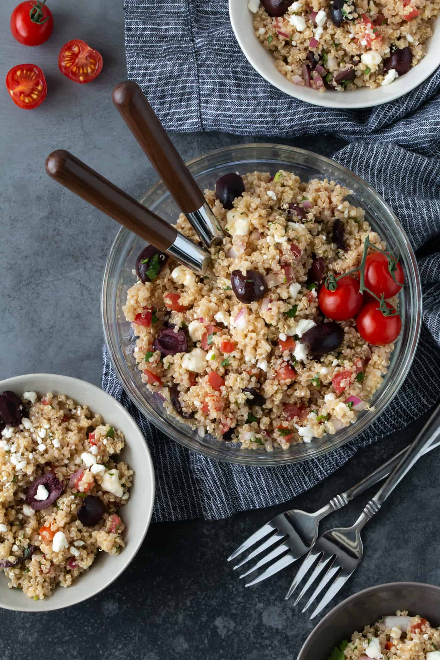 mediterranean quinoa salad in a glass serving bowl with wooden spoons for serving on a dark grey background.
