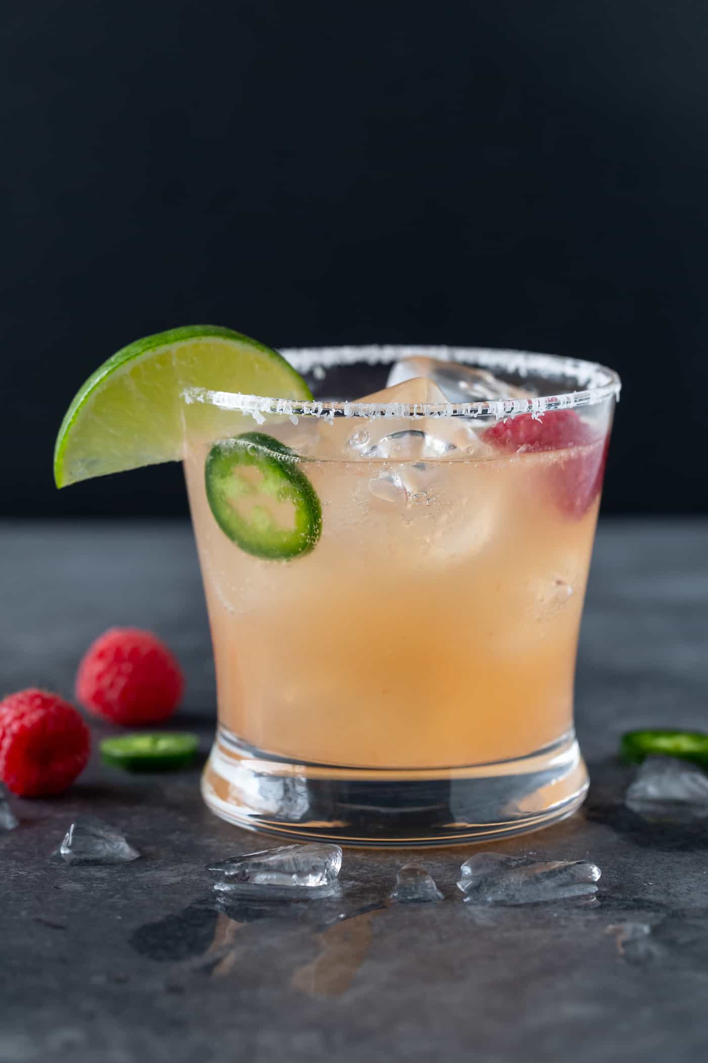 A lowball glass with a salted rim filled with Raspberry Paloma Cocktail and garnished with fresh raspberries, jalapeno slices and a lime wedge.