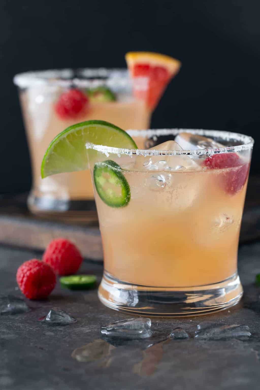 Two lowball glassed rimmed with salt and filled with Raspberry Paloma Cocktails, with lime and grapefruit wedges, fresh raspberries and jalapeno slices.