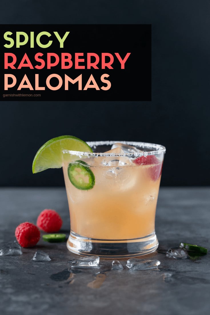 A single lowball glass with a salted rim is filled with Raspberry Paloma Cocktail and garnished with a lime wedge, fresh raspberries and jalapeno slices.