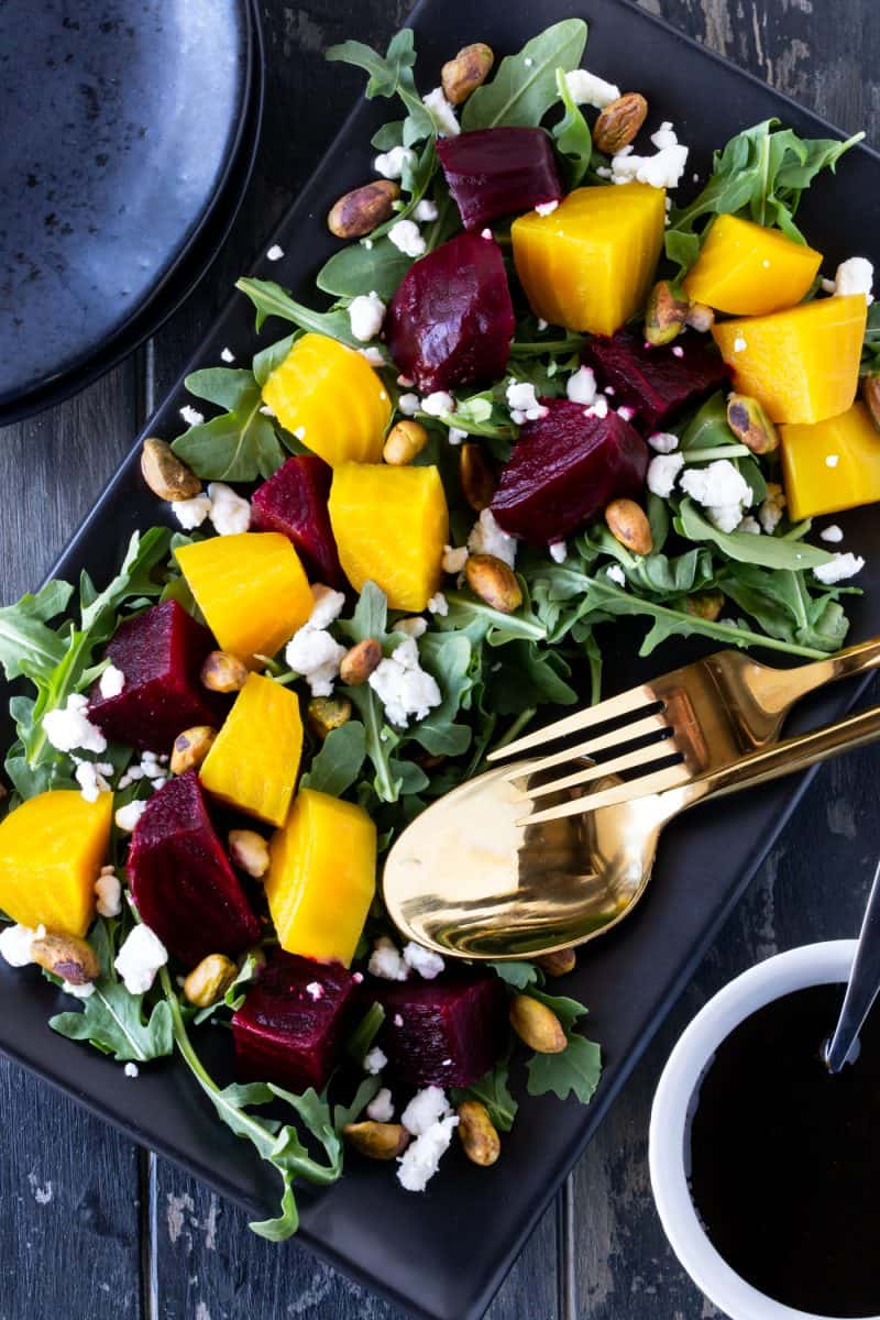 Roasted Beet Salad with Goat Cheese, Arugula and Pistachios on a dark black platter with bowl of balsamic glaze and stack of black plates nearby.