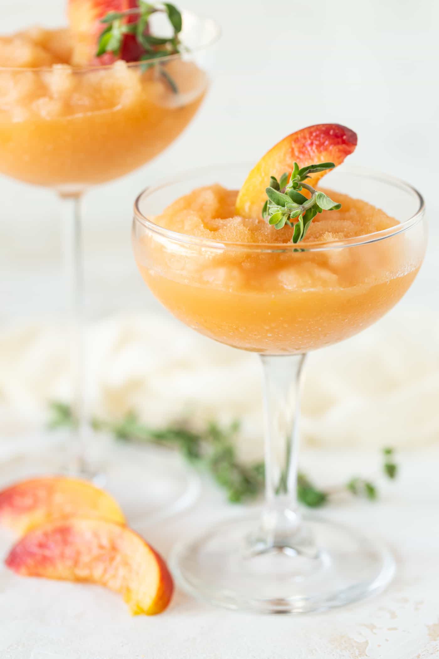 Two coupe cocktail glasses filled with Peach Frosé. Garnished with fresh peach wedges and fresh marjoram.