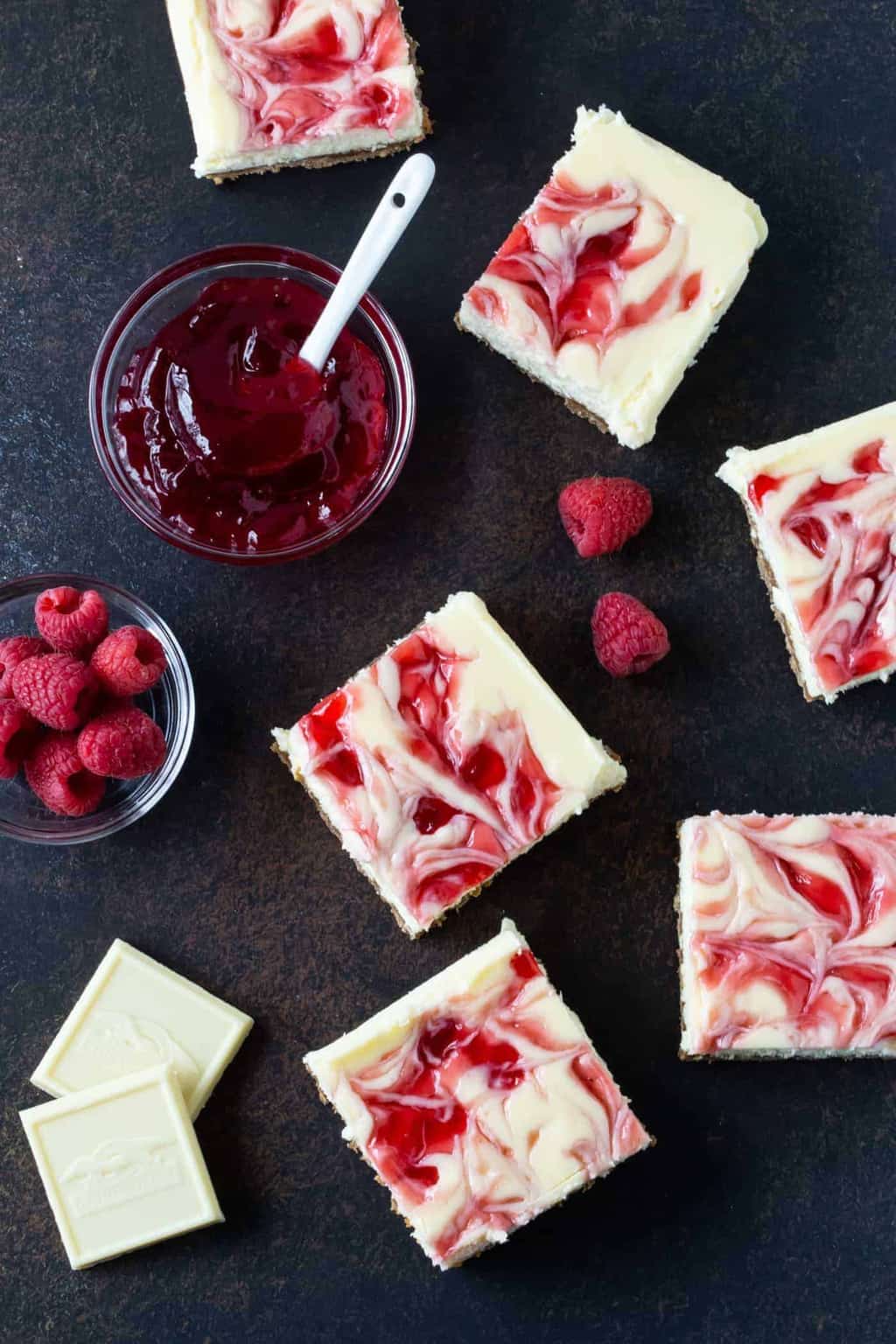  raspberry white chocolate cheesecake bars on a dark board with small bowl of raspberry jam on side.