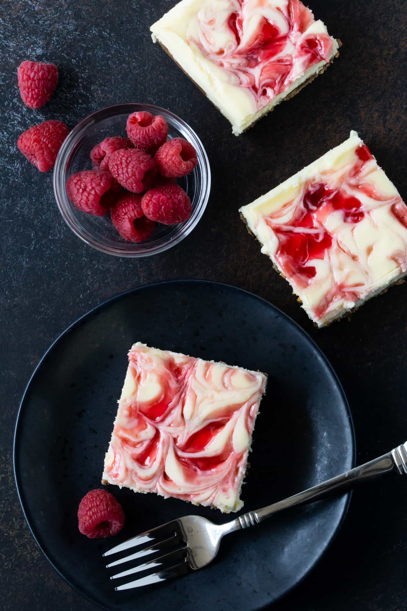 Cut Raspberry White Chocolate Cheesecake Bars on a black plate with fresh raspberries on side.