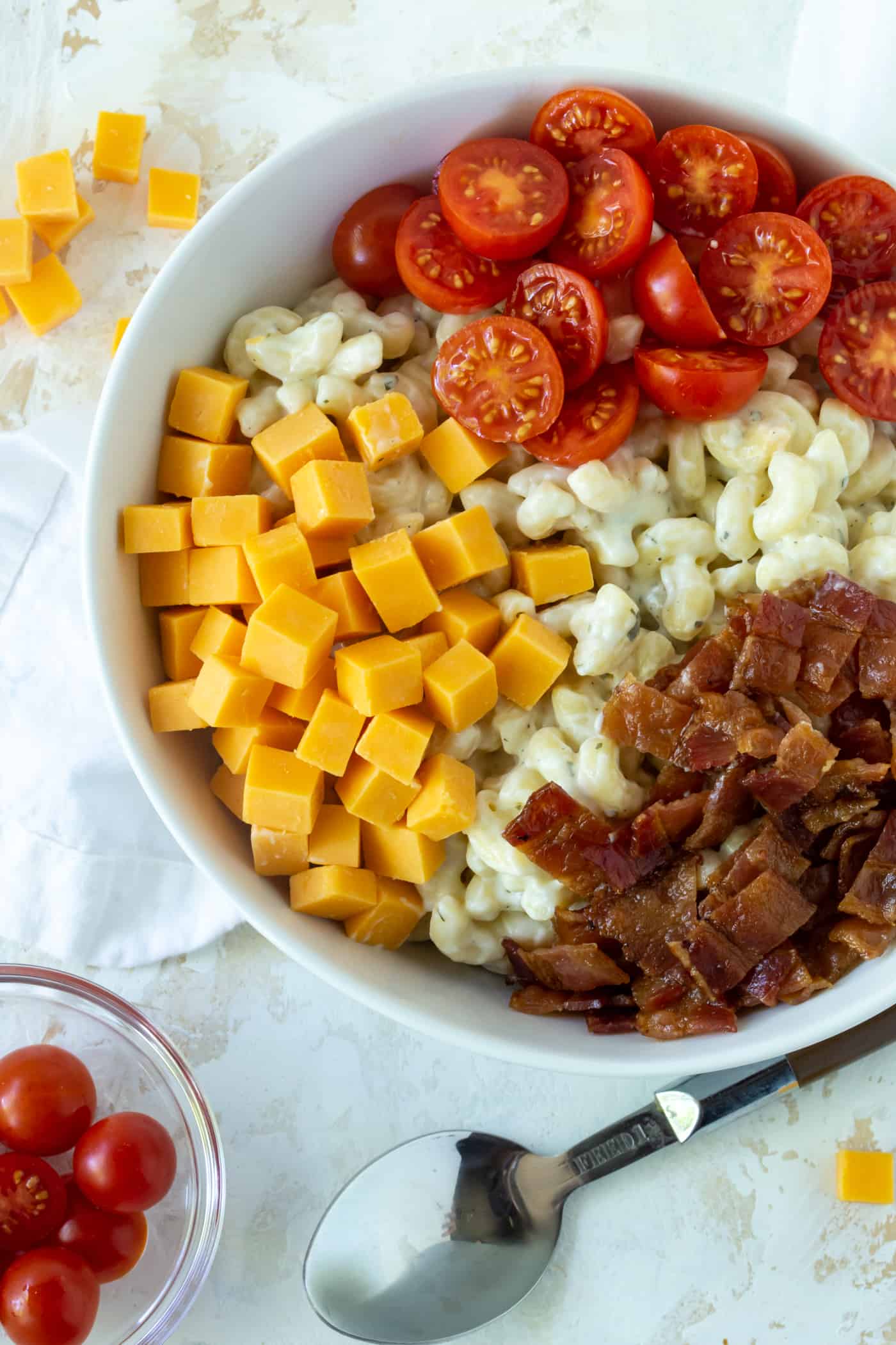 white bowl filled with cooked elbow pasta, cubes yellow cheddar cheese, sliced grape tomatoes and chopped cooked bacon.
