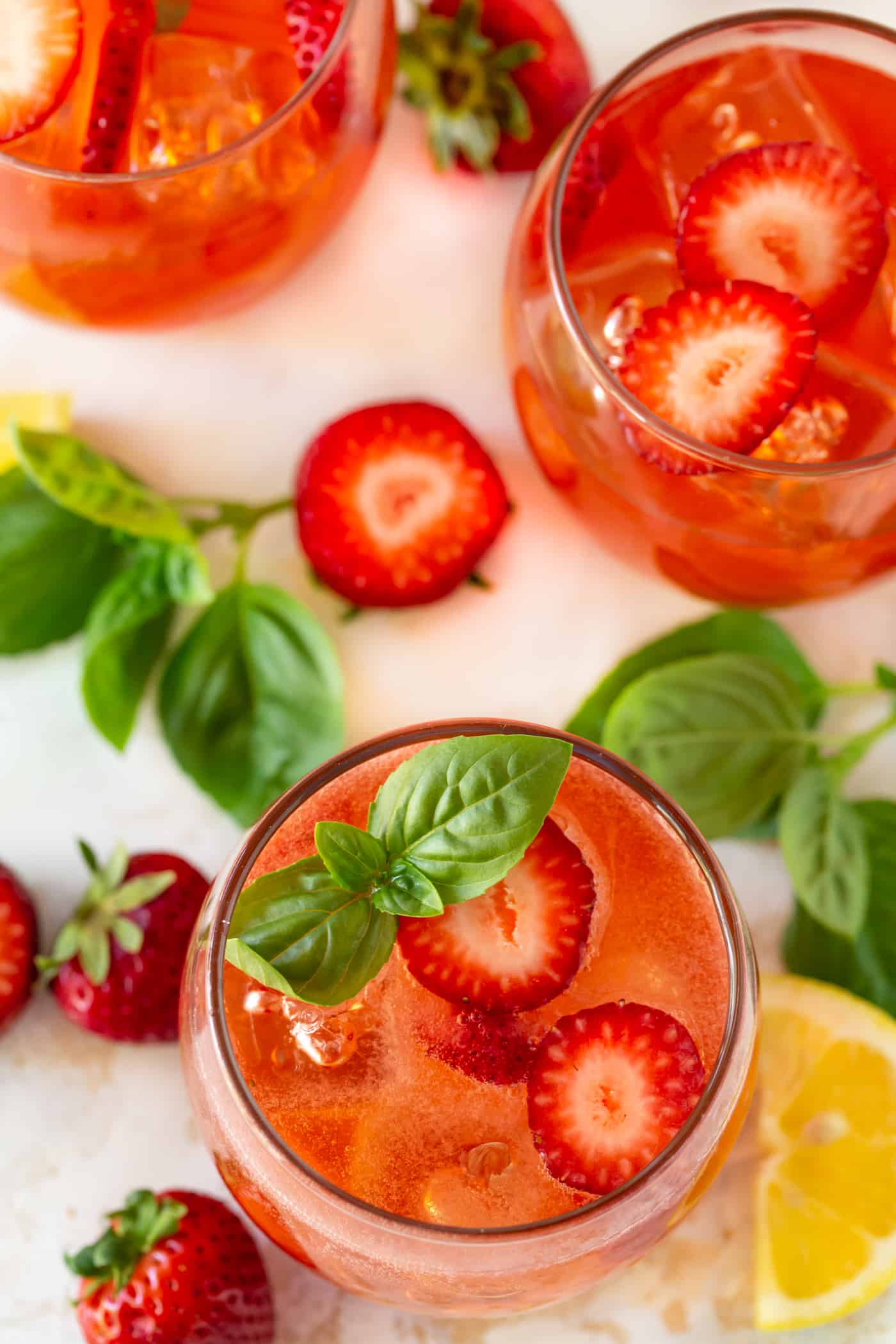 Red colored drink in low ball glass filled with ice. Garnished with fresh berry slices, fresh basil leaves and lemon wedges. 