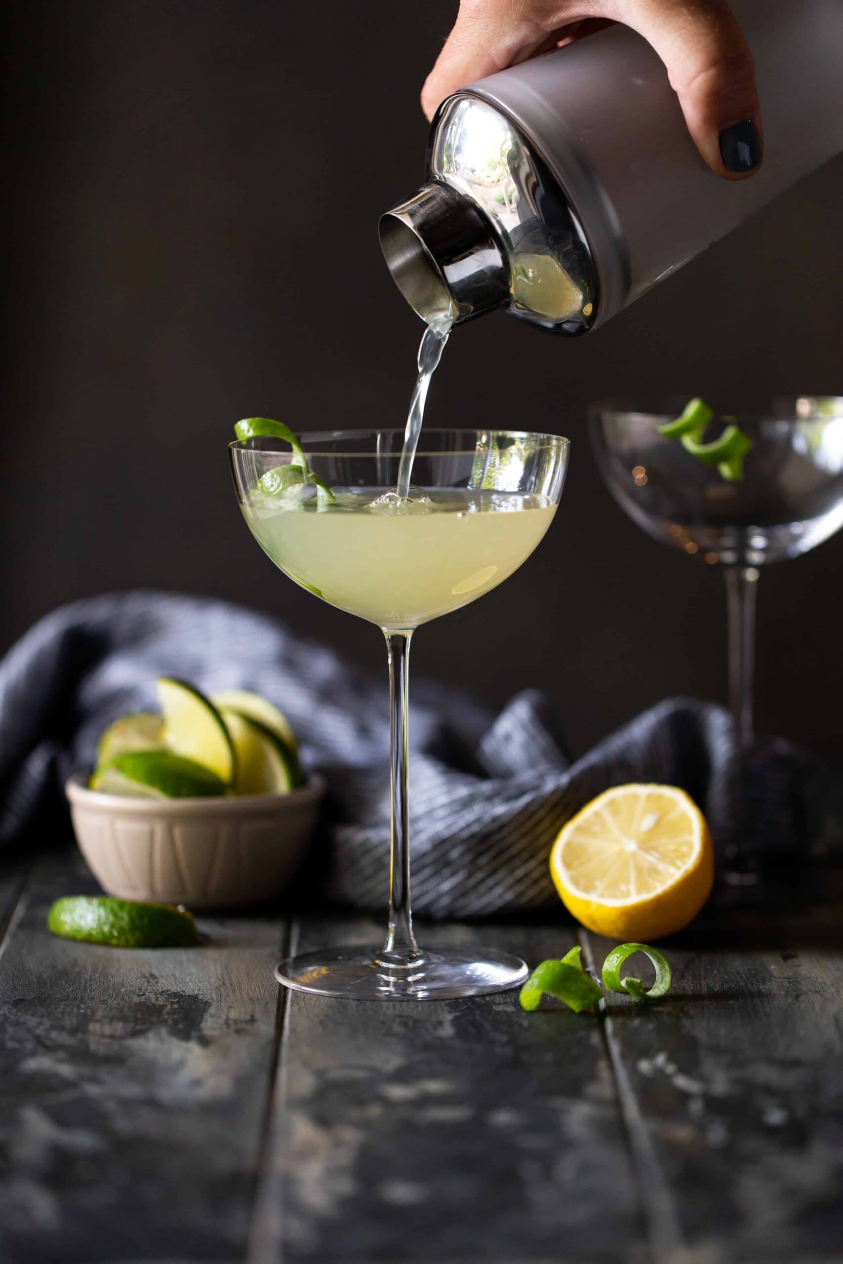  Martini being poured from a shaker into a tall coupe glass with fresh lemon and limes.