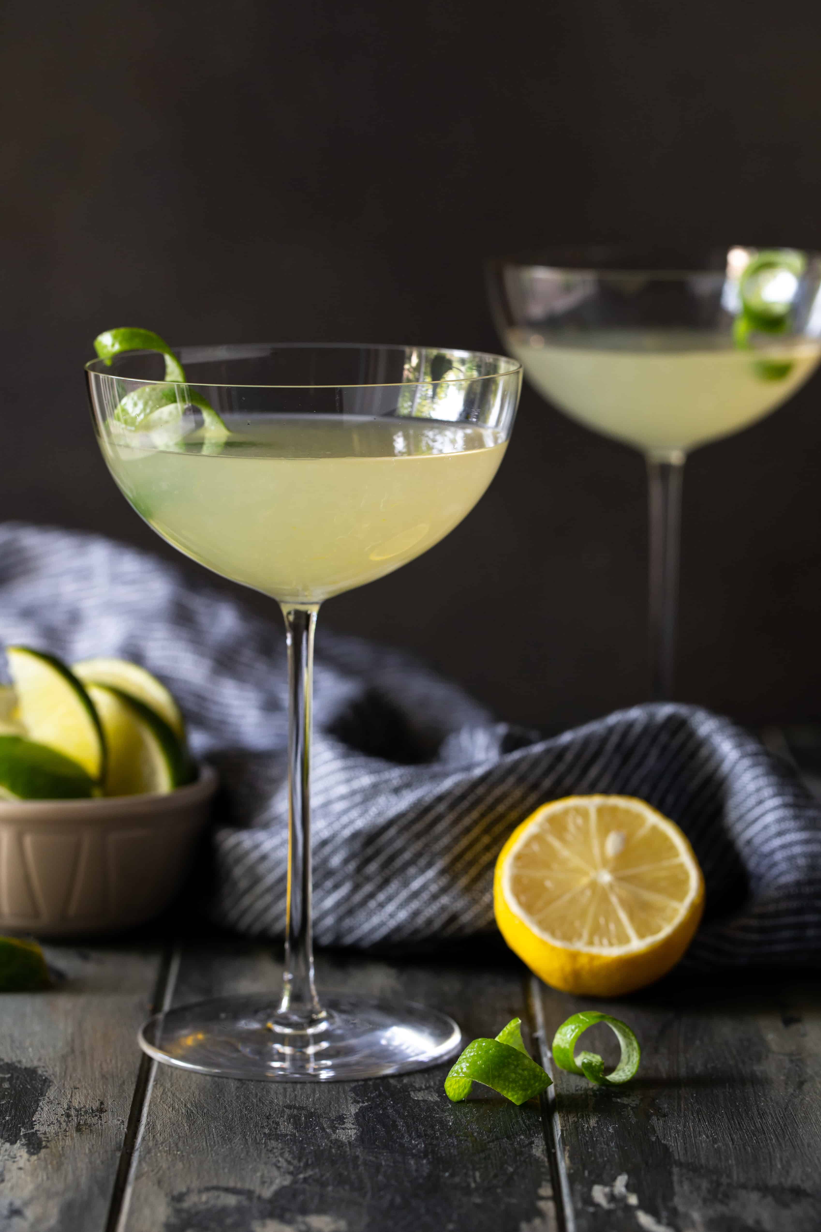 2 martinis in tall coupe glasses with lime twists for garnish on a dark background.