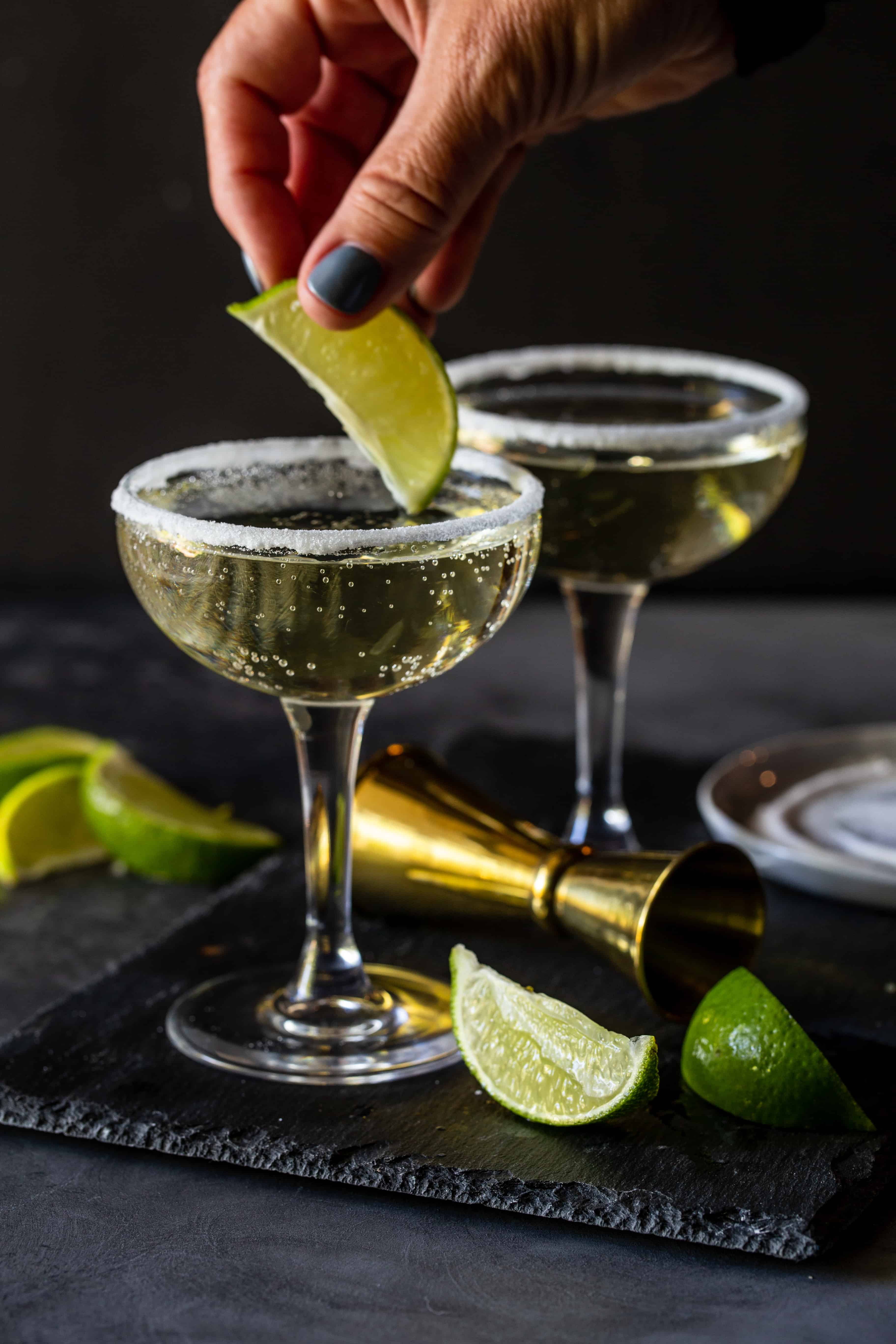 Lime wedge being dunked in coupe glasses that are rimmed with sugar and filled and extra lime wedges for garnishes.