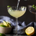 Martini shaker pouring drink into large coupe glass with lime twist.