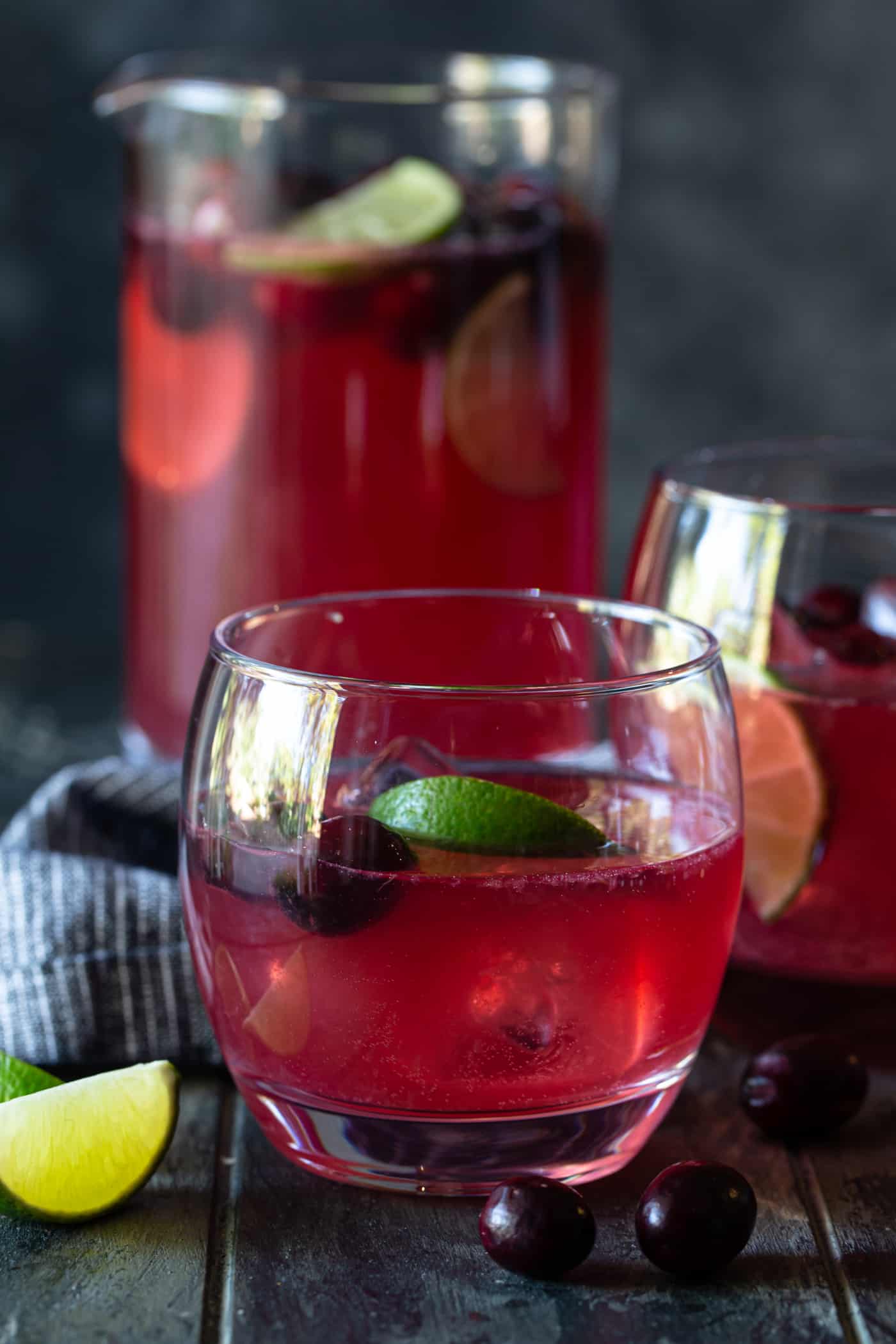 A pitcher and low ball glass filled with Punch,  with fresh lime wedges and fresh cranberries.