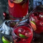 Pitcher filled with Cranberry Vodka Party punch, with fresh cranberries and lime slices and surrounded by lowball glasses filled with punch.