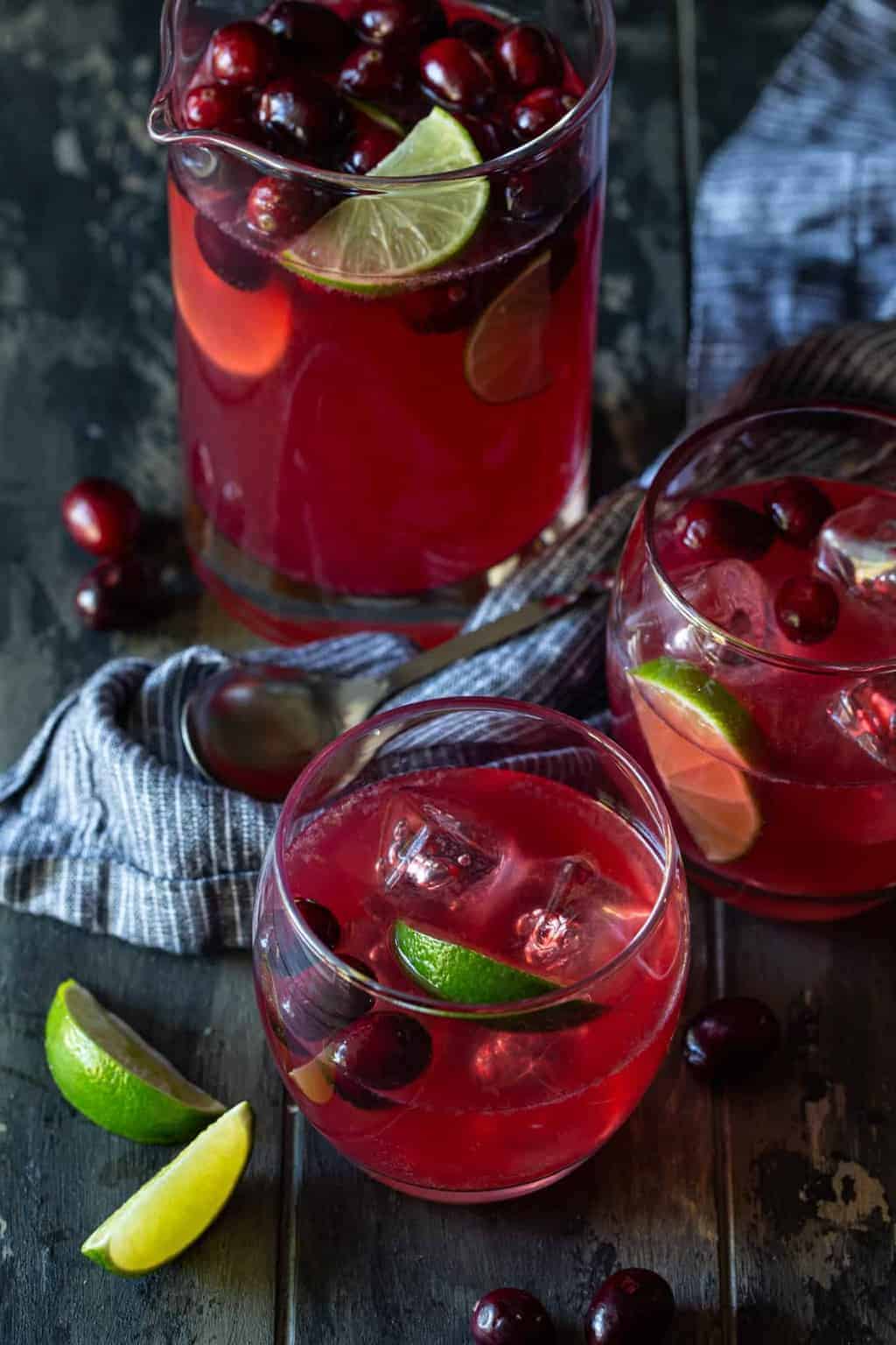 Pitcher filled with Cranberry Vodka Party punch, with fresh cranberries and lime slices and surrounded by lowball glasses filled with punch.