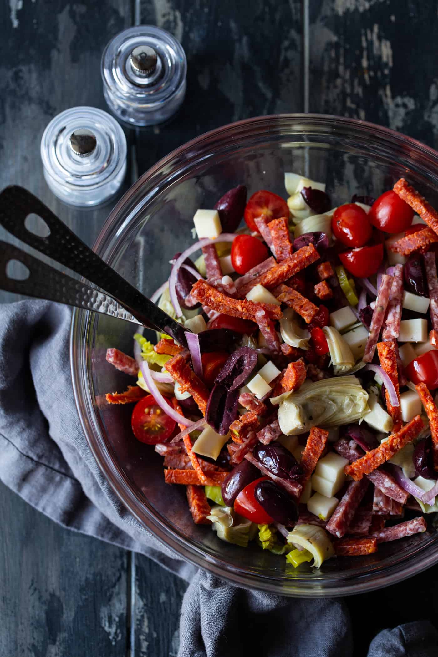 Salad in a large glass bowl with spoons for tossing and salt and pepper shaker on the side.