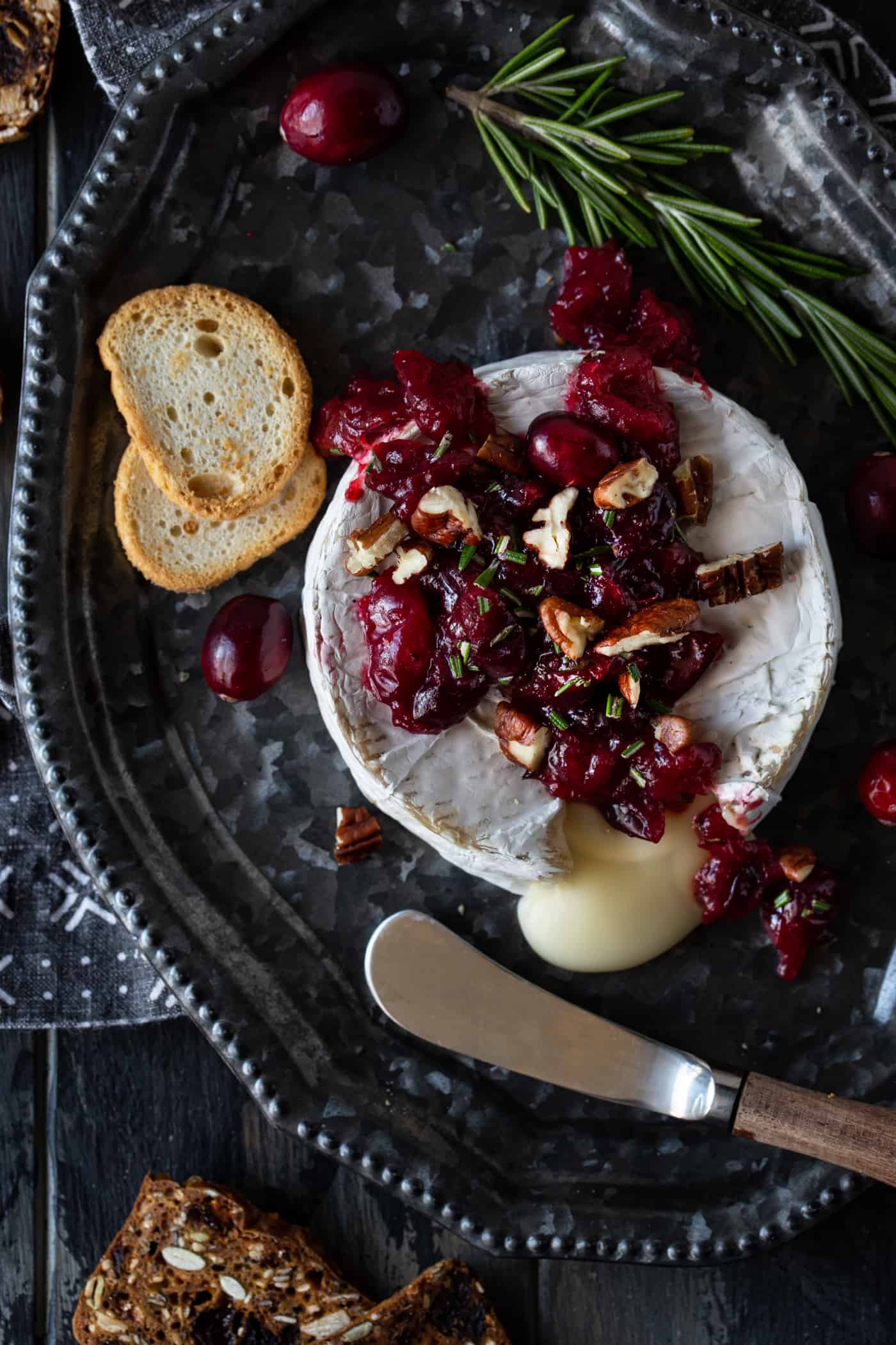 A wheel of warm brie with Cranberries and Pecans on a silver tray with a spreader and mini toasts.