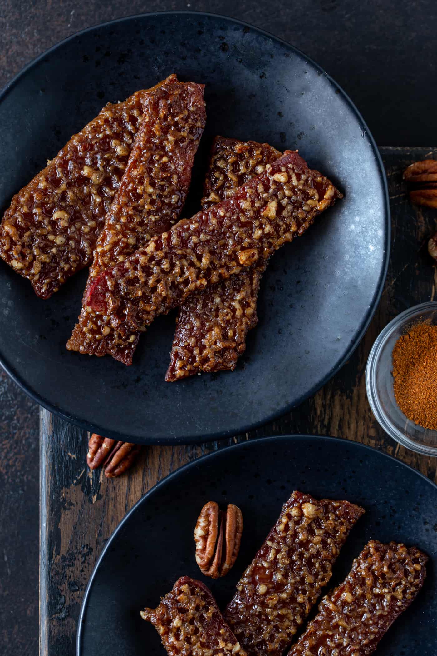 Candied Bacon on 2 black plates with pecan and cayenne pepper for garnish.