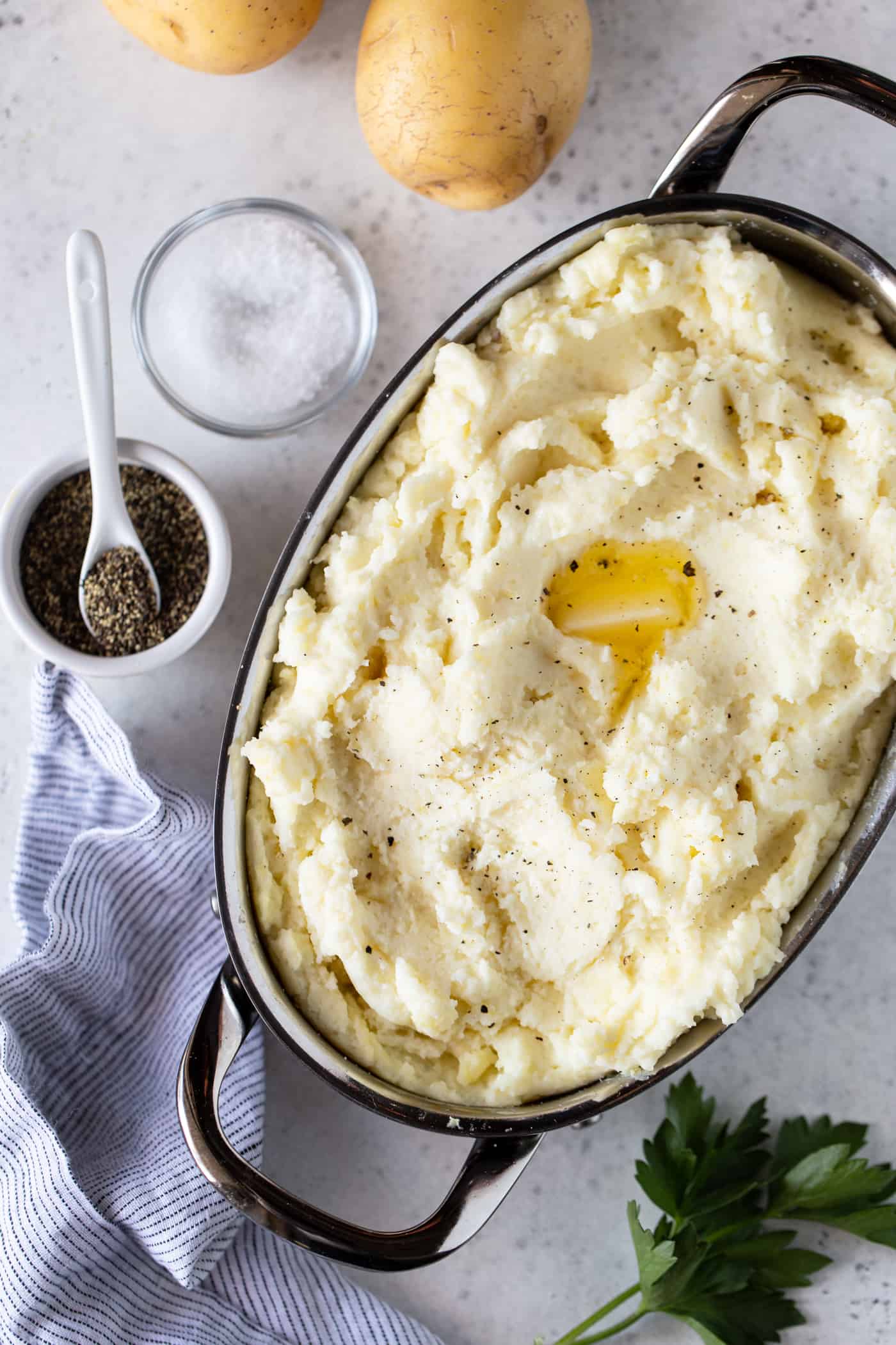 Mashed potato in a stainless steel oval baking dish with melted butter, salt and pepper on top.