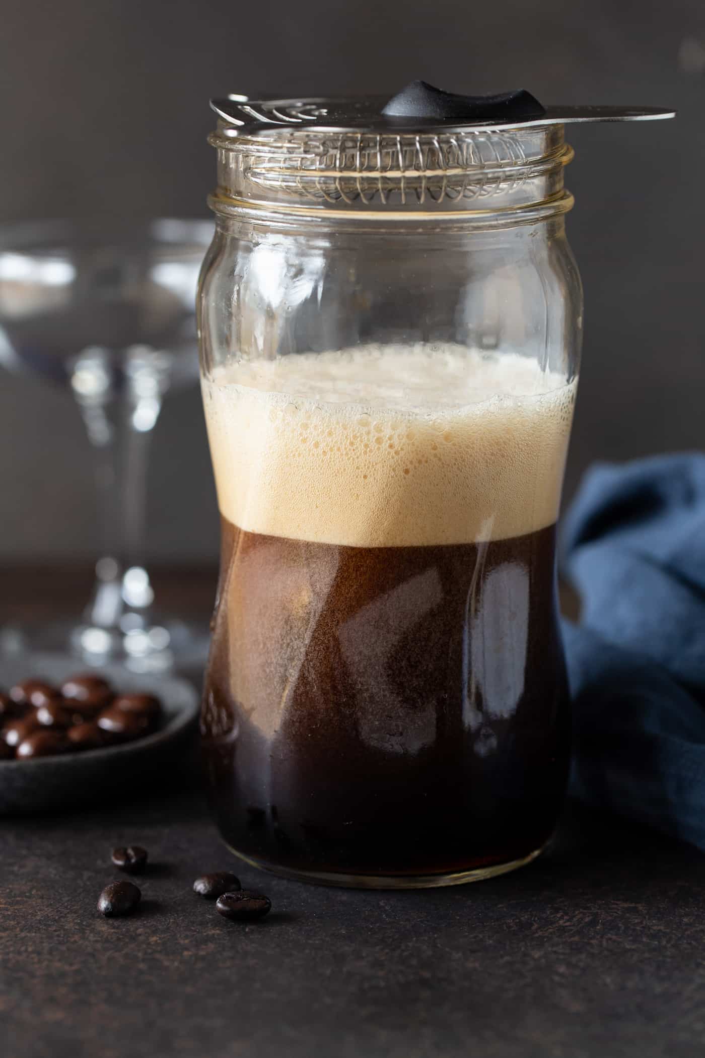 Mason jar filled with frothy espresso martini and strainer on top with an empty martini glass in the background.