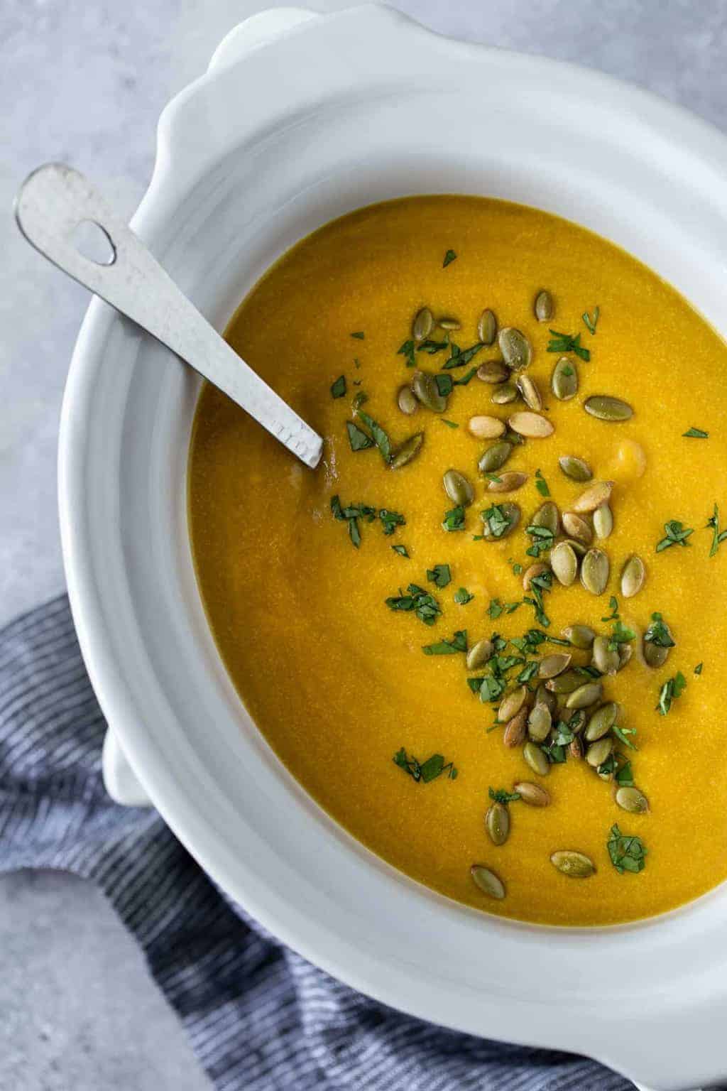 White slow cooker filled with Butternut Squash Soup. Garnished with chopped fresh cilantro and pepitas.