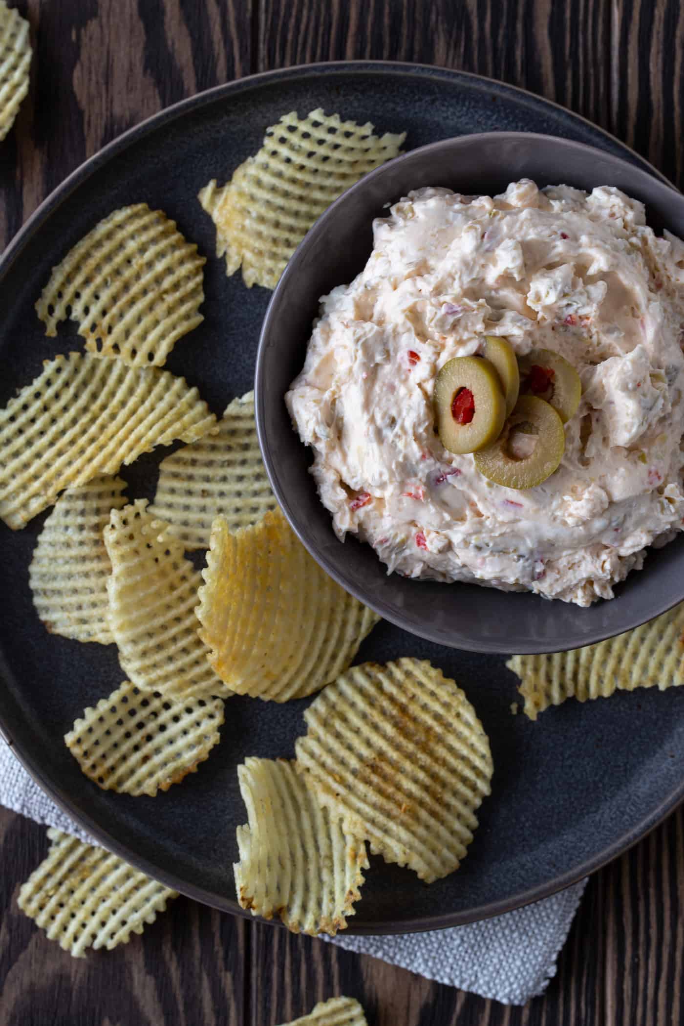 Wooden bowl filled with Green Olive Dip and garnished with sliced green olives and crackers. Green olive dip on a plate, with sliced olives and chips.