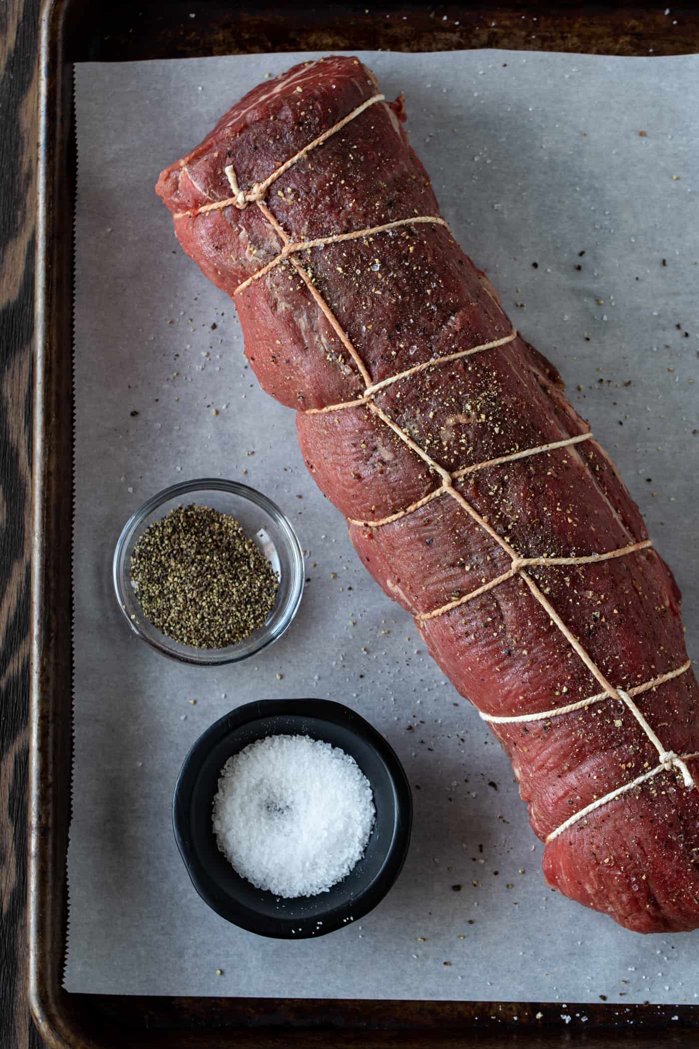 Tied roast of beef on a baking sheet with salt and pepper on side.