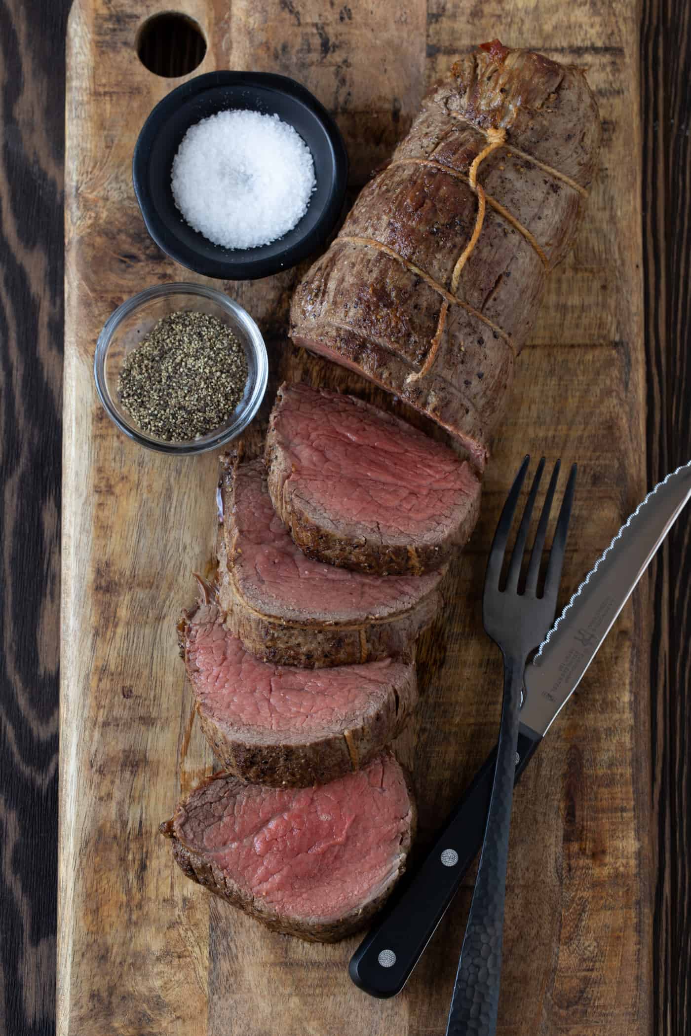 Sliced beef tenderloin on a brown cutting board with salt and pepper bowls on side. 