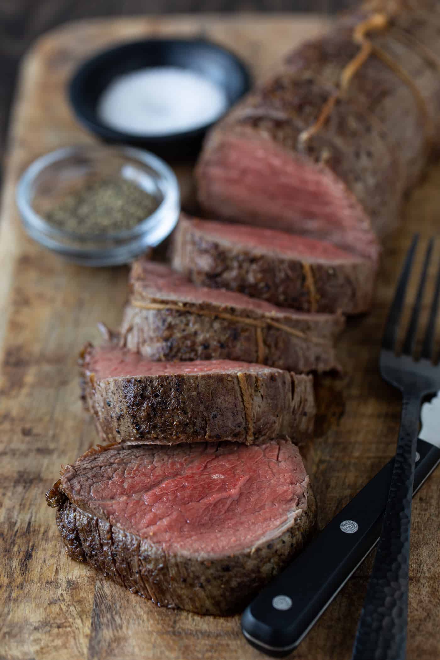 Sliced beef beef tenderloin on cutting board with steak knife and fork.