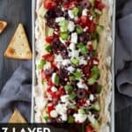 Layered Greek Dip on a white platter. Garnished with pita chips. 