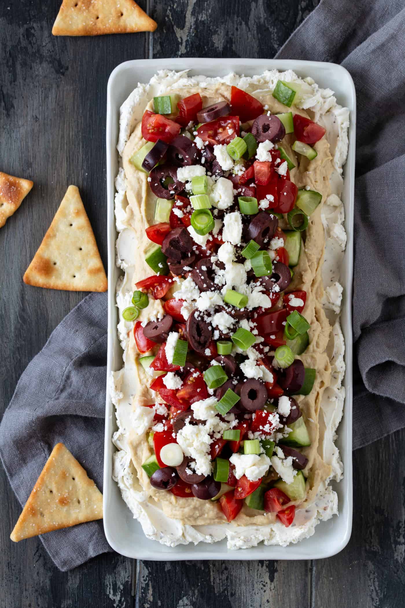 A white platter filled with Layered Greek Dip and garnished with pita wedges.