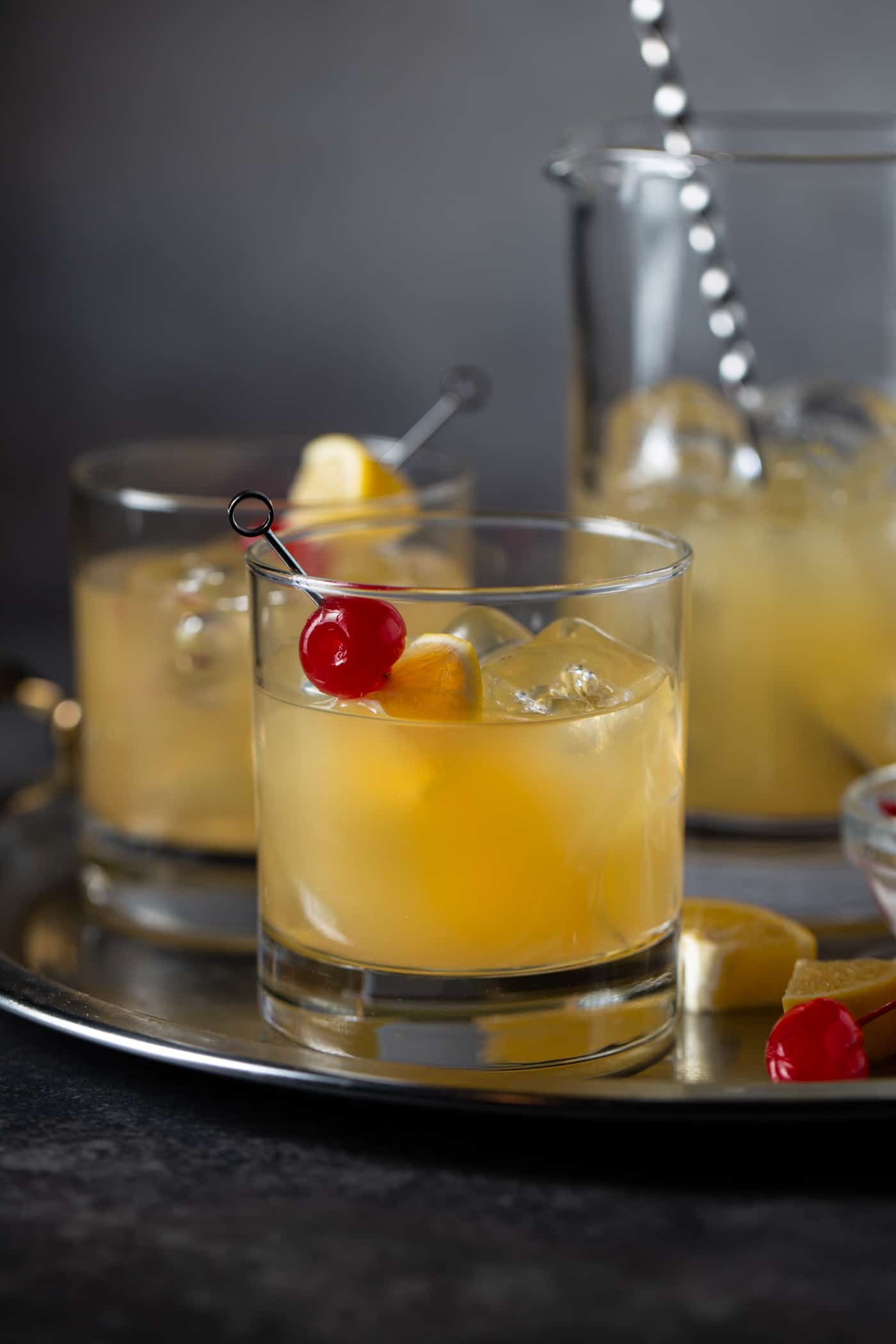  Drinks in lowball glasses filled with ice on a silver tray. A pitcher of cocktails is in the background.