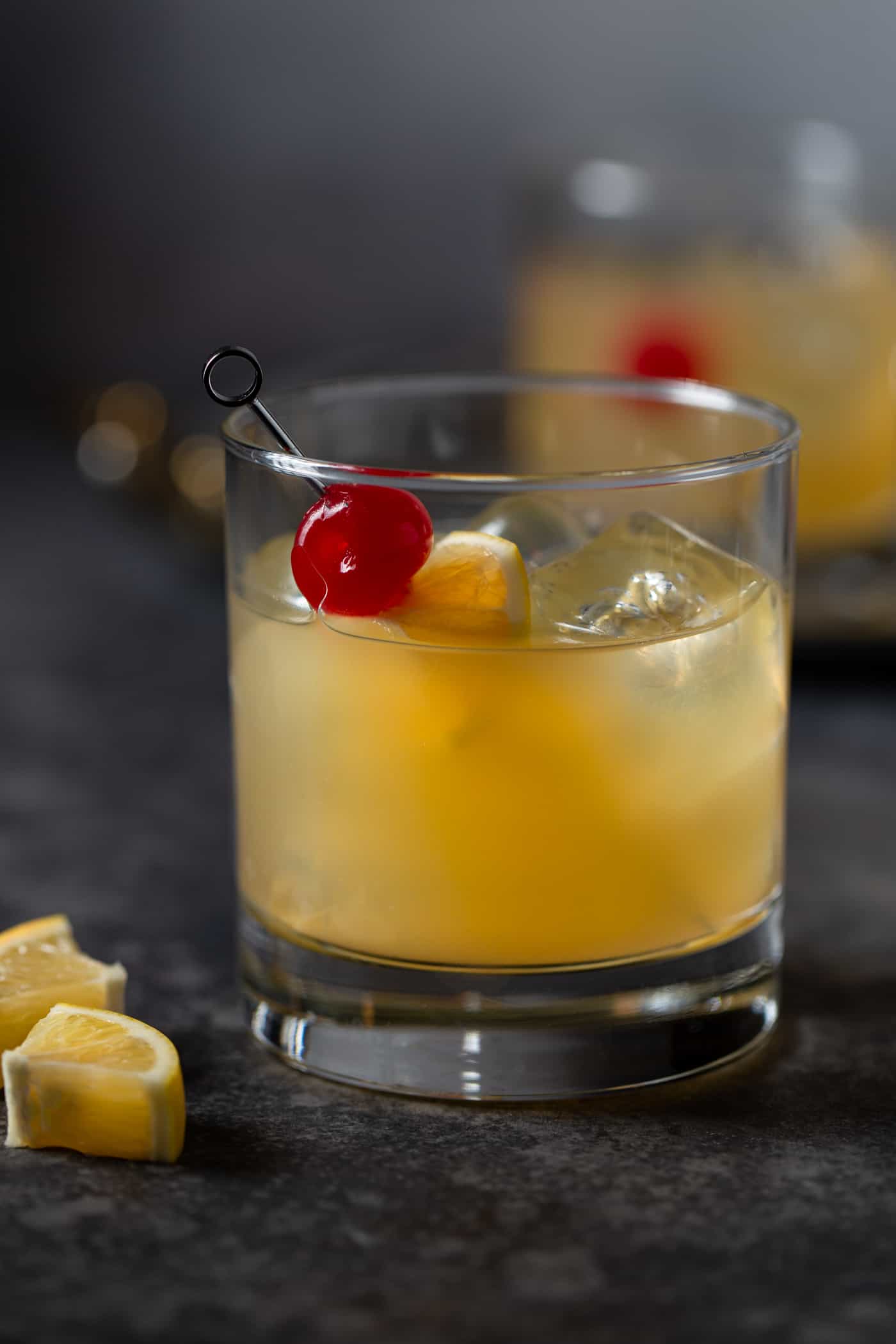 A cocktail in a low ball glass filled with ice and garnished with maraschino cherries and Meyer lemon slices.