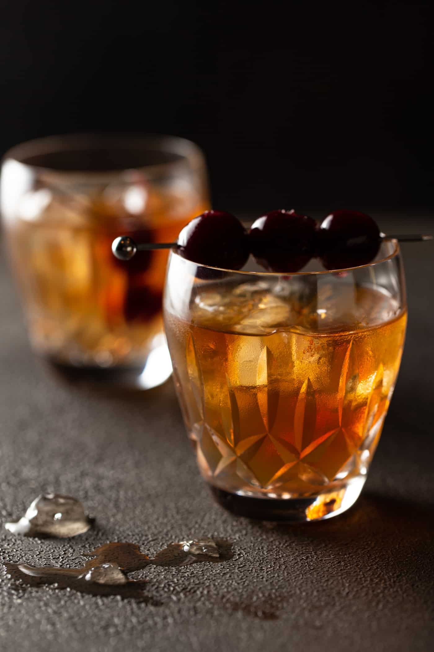 2 crystal glasses filled with ice, bourbon and a skewer of cherries for garnish on a dark background.