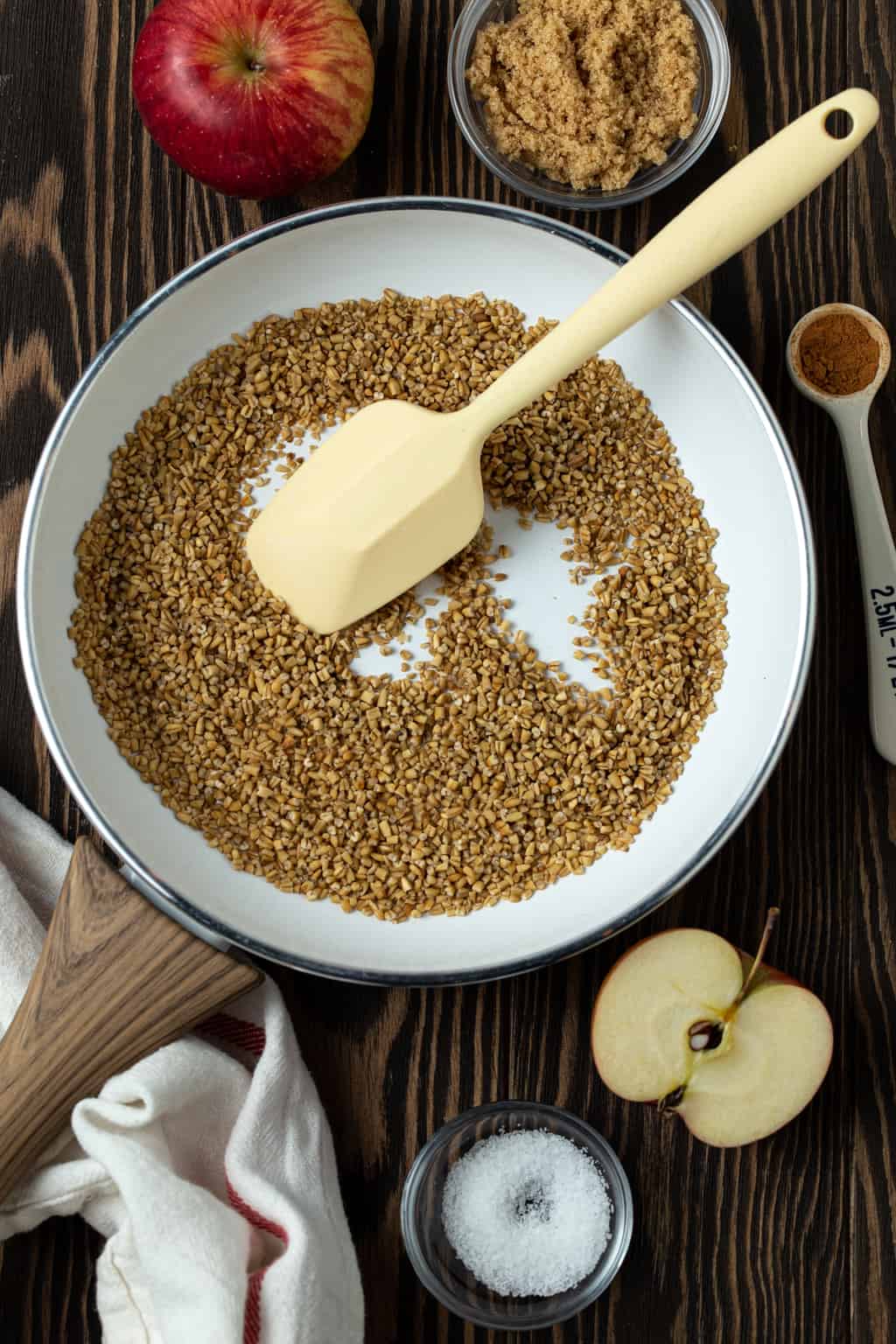 Toasted Steel Cut oats in a white frying pan with sliced apples and cinnamon sticks.