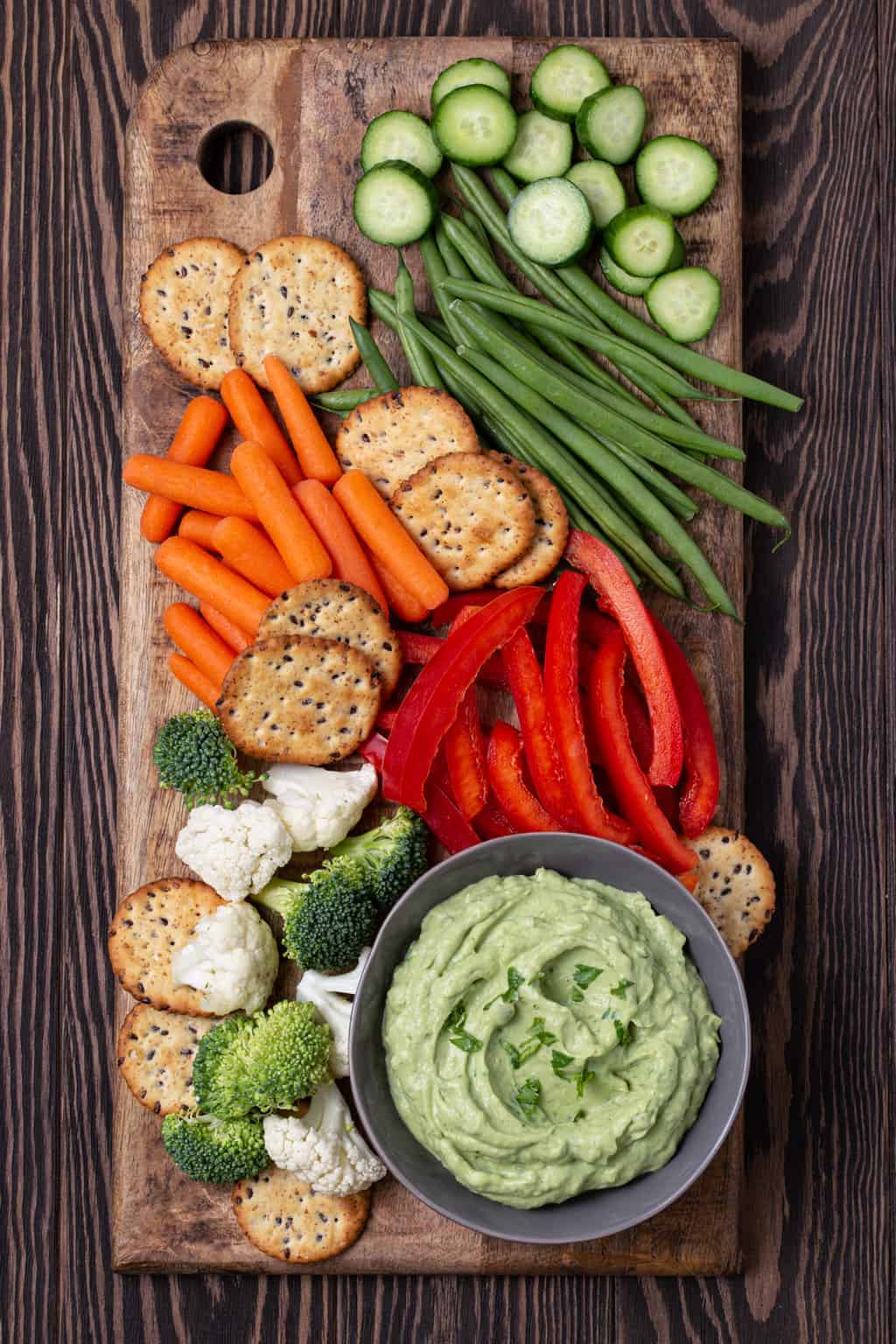 Wooden cutting board filled with vegetables, crackers and a bowl filled with a healthy Avocado Dip that is made with Greek yogurt