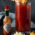 High Ball glass filled with Bloody Mary and garnished with skewer filled with shrimp, cheese and olive. Tabasco bottle and bowl of horseradish for garnish.