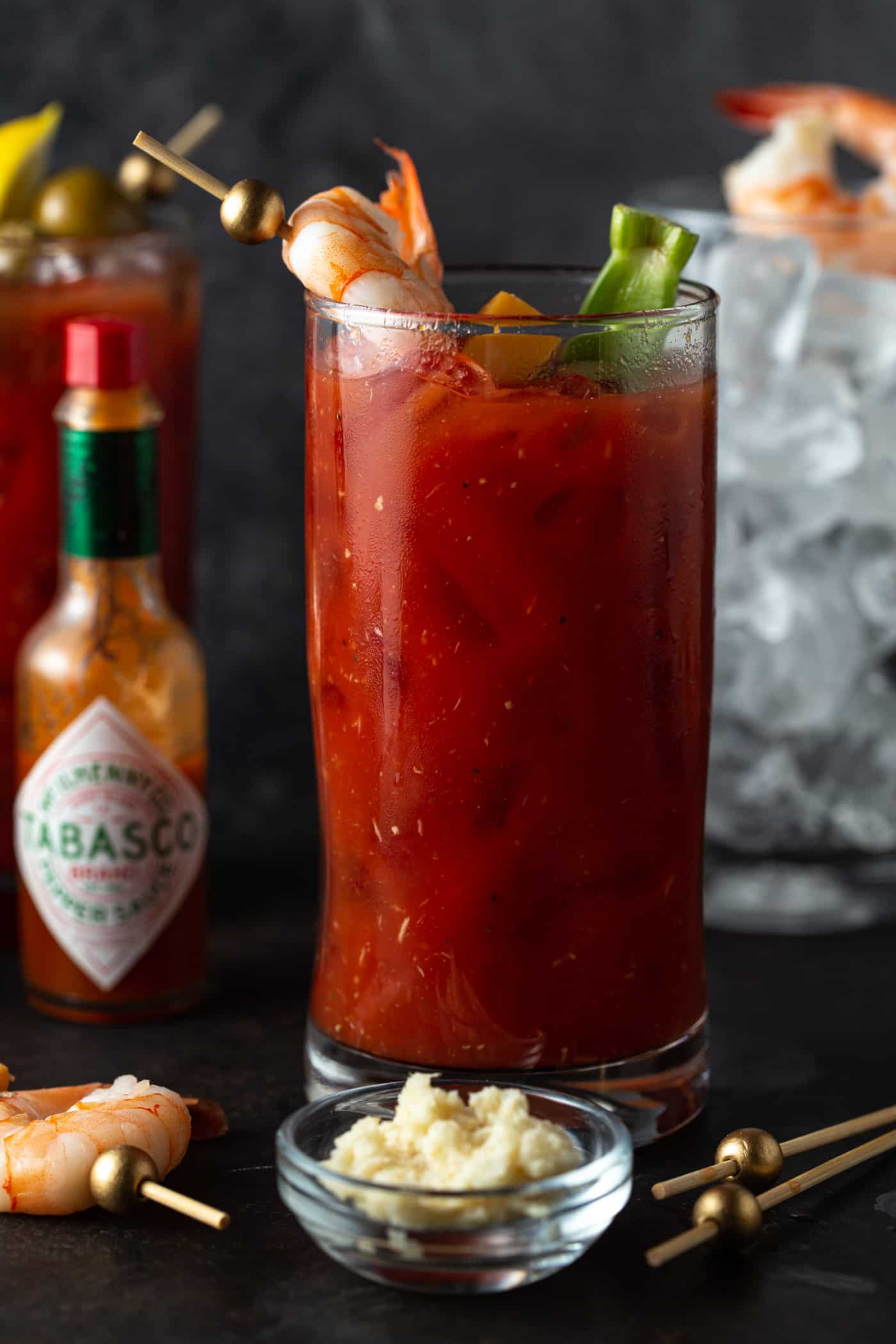 High Ball filled with drink and garnished with skewer of olives and shrimp. Empty glass filled with ice in the background on a dark surface.