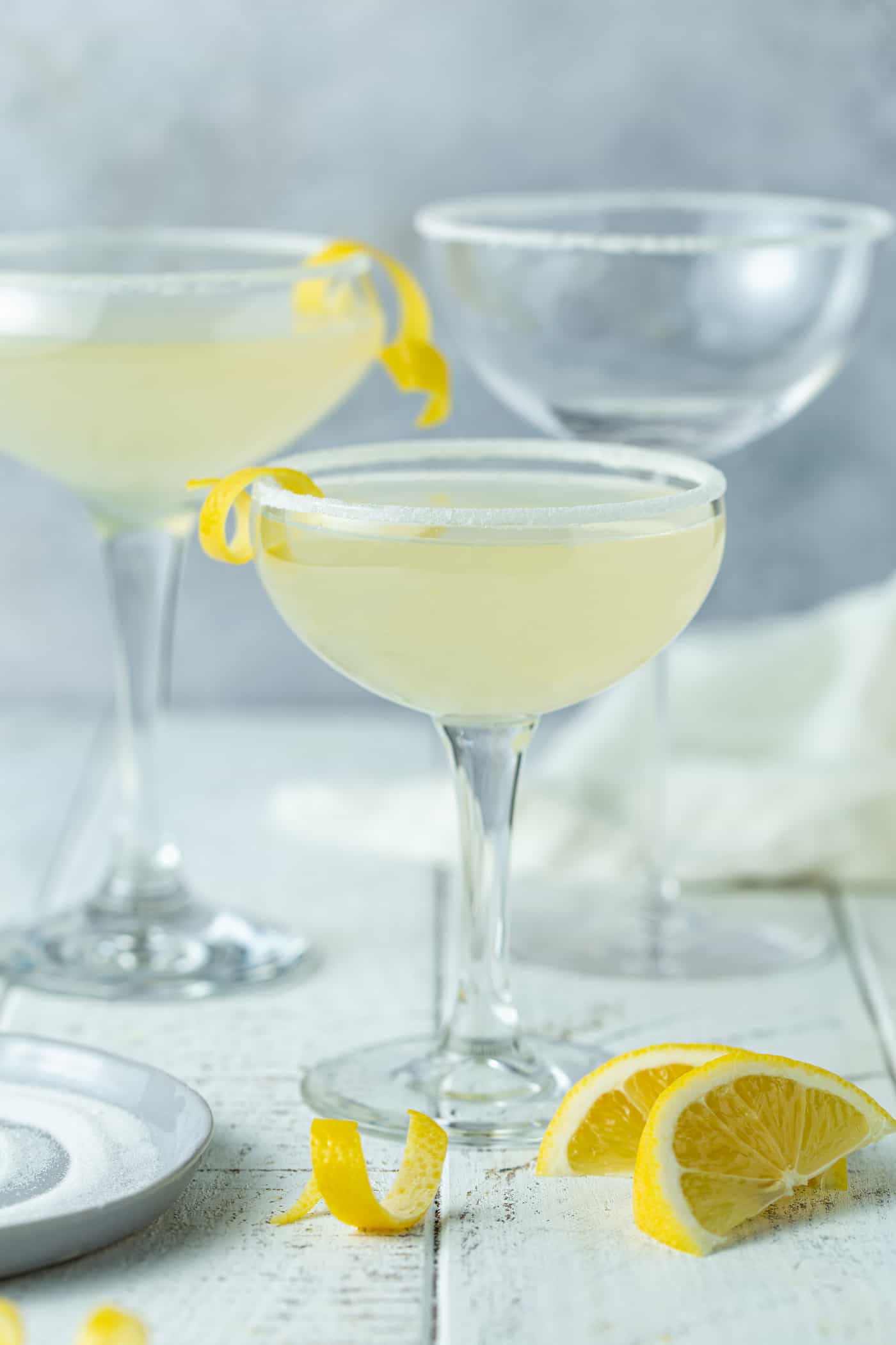 3 martini glasses on a gray background filled with lemon drop martinis and garnished with lemon twists.