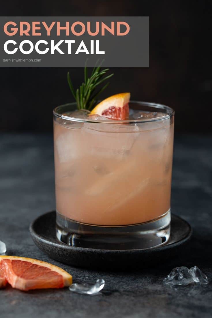 Single low ball glass on a dark gray background filled with ice, grapefruit juice and gin. it is garnished with a fresh wedge of grapefruit and fresh rosemary.