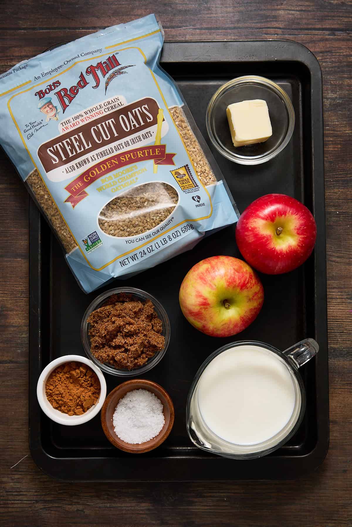 A bag of steel cut oats, butterm apples, cinnamon, brown sugar, salt and milk on a black tray.