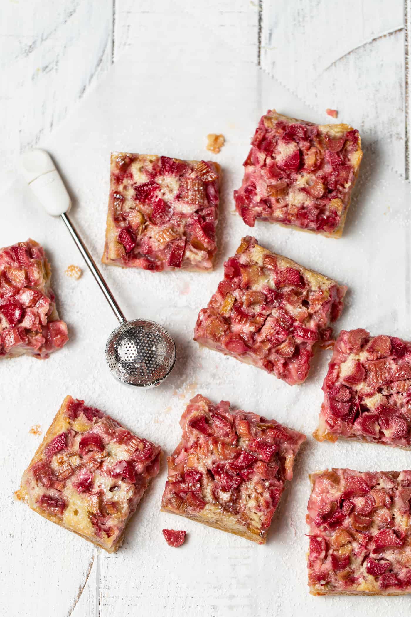 rhubarb bars cut into squares and scattered on a white background dusted with powder sugar.