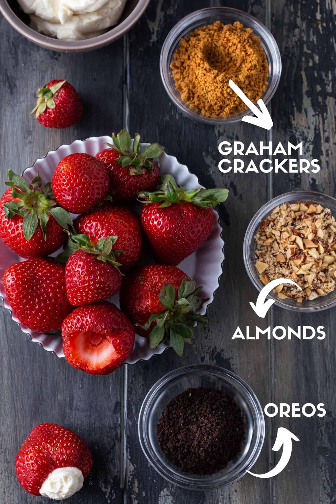 Small bowls of crushed Oreos, graham crackers and almonds next to strawberries and cheesecake filling. 