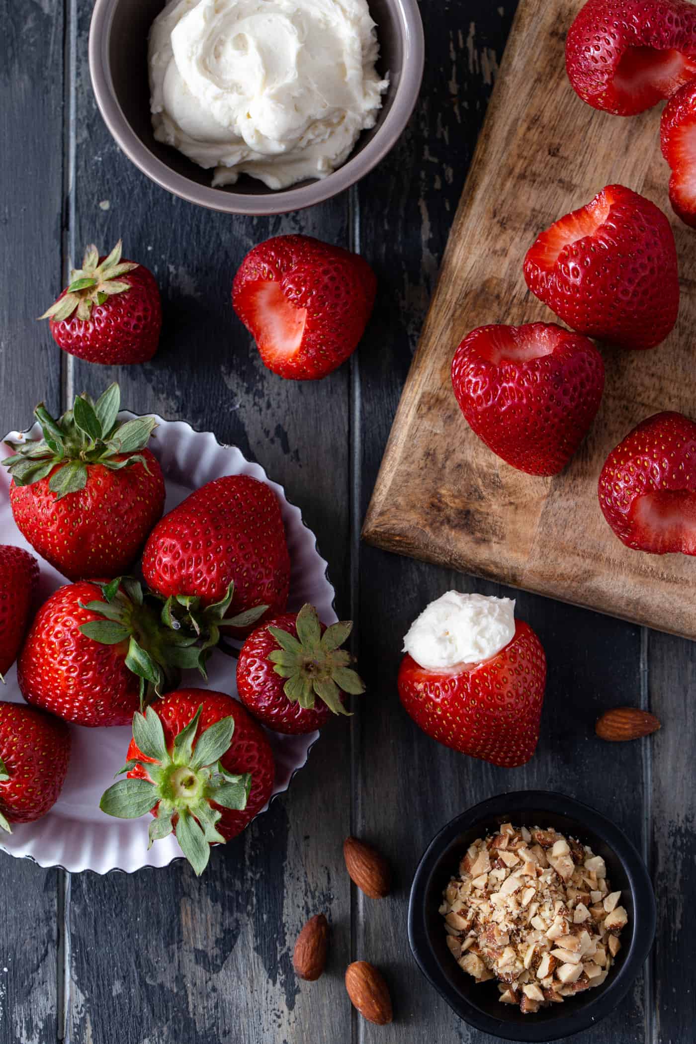 Strawberries on a wood cutting board with a bowl of cheesecake filling and a bowl of crushed almonds. 