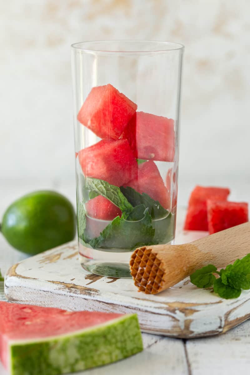 A collins glass filled with the ingredients for a watermelon mojito: watermelon cubes, mint leaves, lime juice and sugar.