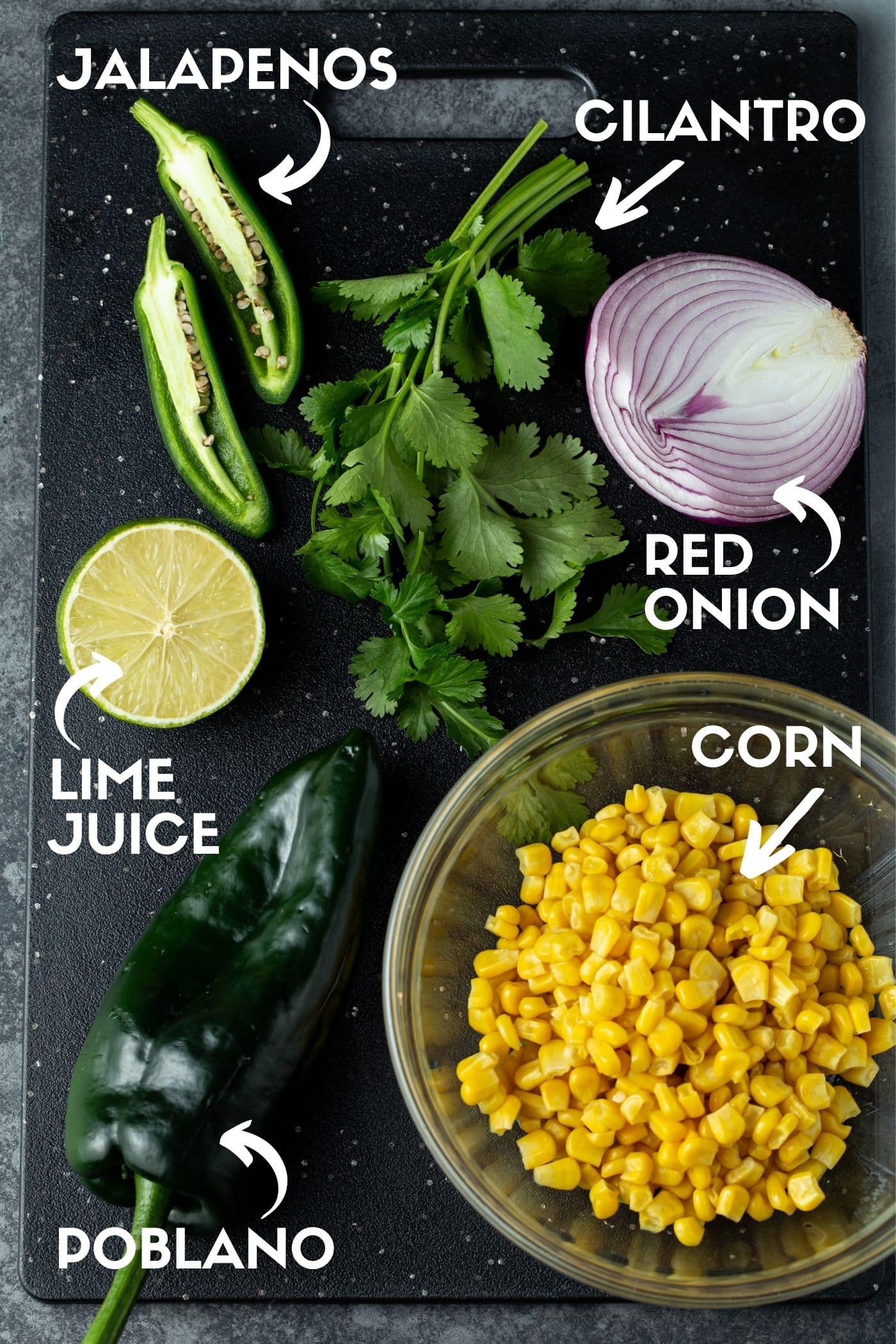 Ingredients for corn salsa on a black cutting board, including corn, cilantro, jalapeños, red onion, poblano peppers and half of a lime. 