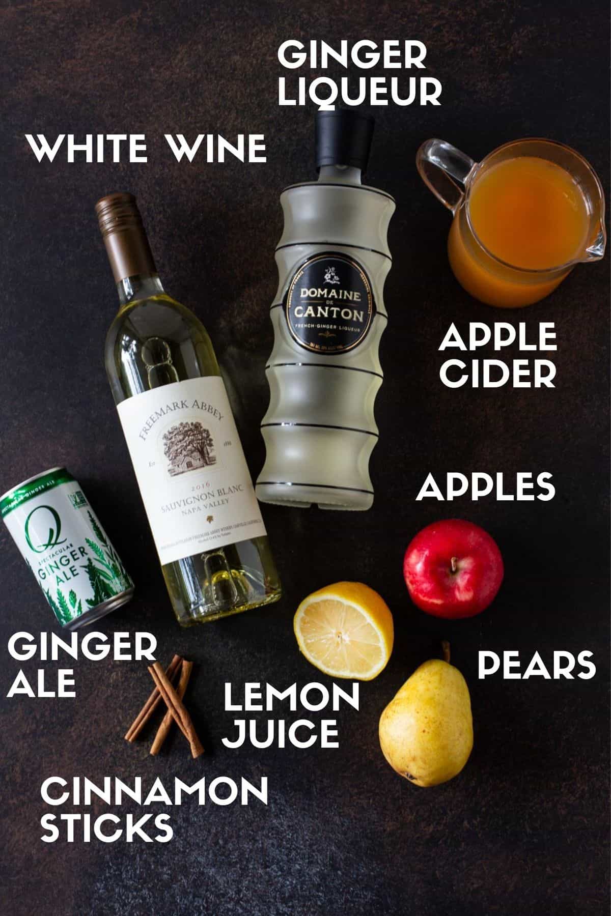 Ingredients for Apple Cider Sangria on a dark board, including apple cider, ginger liquer, apples, pears, lemon juice, white wine and cinnamon sticks. 